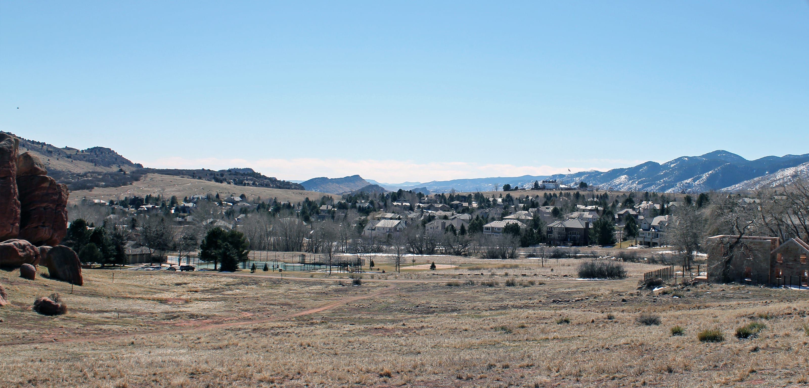 Dry grass and red sandstone outcrops in Ken Caryl, a neighborhood of pitched roofs among bare trees, with the snow-streaked Front Range behind it